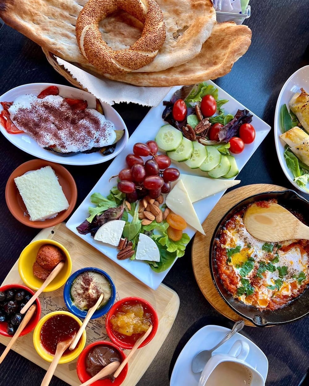 An overhead spread of Afghan breakfast: fresh naan and sesame bread, borani with sumac, white cheese, olives, jams, a herb salad and eggs baked in tomato.