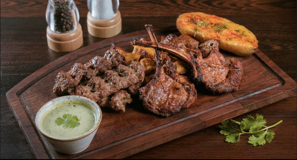A wooden board of grilled lamb chops and minced-beef kabob with garlic naan and a bowl of green chutney.