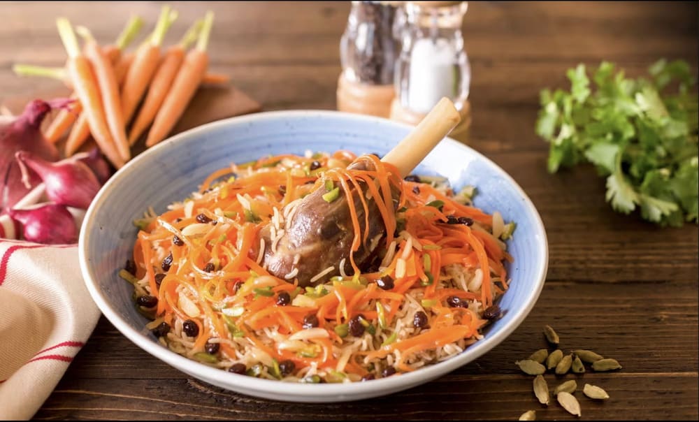 Kabuli palaw — a lamb shank buried in long-grain rice with shredded carrot, raisins and pistachio, in a blue-rimmed bowl.